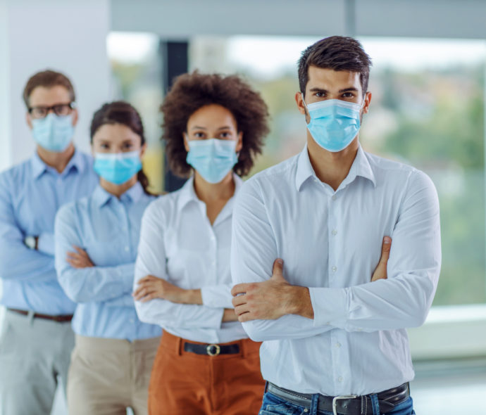 Multicultural group of business people with face masks on standi Multicultural group of business people with face masks on standing in office with arms crossed and looking at camera. Selective focus on man in foreground.